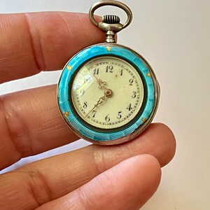 May include: A round pocket watch featuring a bright turquoise guilloch&eacute; enamel bezel with small gold star accents. The watch face is off-white with black Arabic numerals and ornate gold hands. A silver-toned winding crown sits at the top.