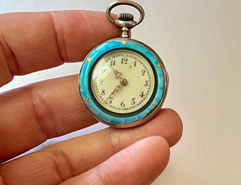 May include: A round pocket watch featuring a bright turquoise guilloch&eacute; enamel bezel with small gold star accents. The watch face is off-white with black Arabic numerals and ornate gold hands. A silver-toned winding crown sits at the top.