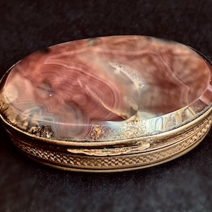 May include: An oval-shaped, gold-toned trinket box with a hinged lid. The lid is made of a translucent, pink and white agate stone with swirling patterns. The box has a decorative, textured border.