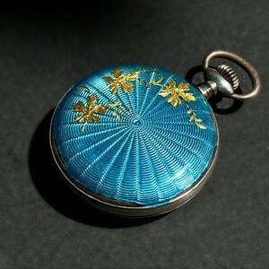 May include: A round pocket watch featuring a vibrant turquoise blue guilloch&eacute; enamel finish with a radiating sunburst pattern. The surface is decorated with delicate gold-toned leaf motifs. It includes a metallic winding crown and a circular loop.