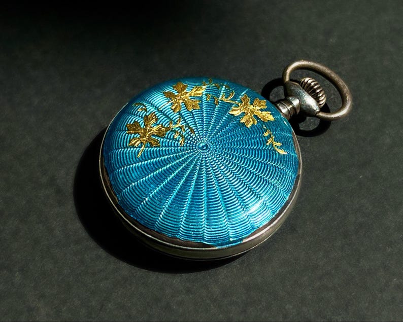 May include: A round pocket watch featuring a vibrant turquoise blue guilloch&eacute; enamel finish with a radiating sunburst pattern. The surface is decorated with delicate gold-toned leaf motifs. It includes a metallic winding crown and a circular loop.