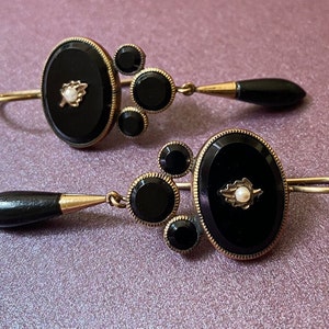 May include: A pair of black and gold earrings with a teardrop-shaped black stone dangle. Each earring features a round black stone with a smaller black stone on either side. The center stone is surrounded by a gold setting with a small pearl in the center.