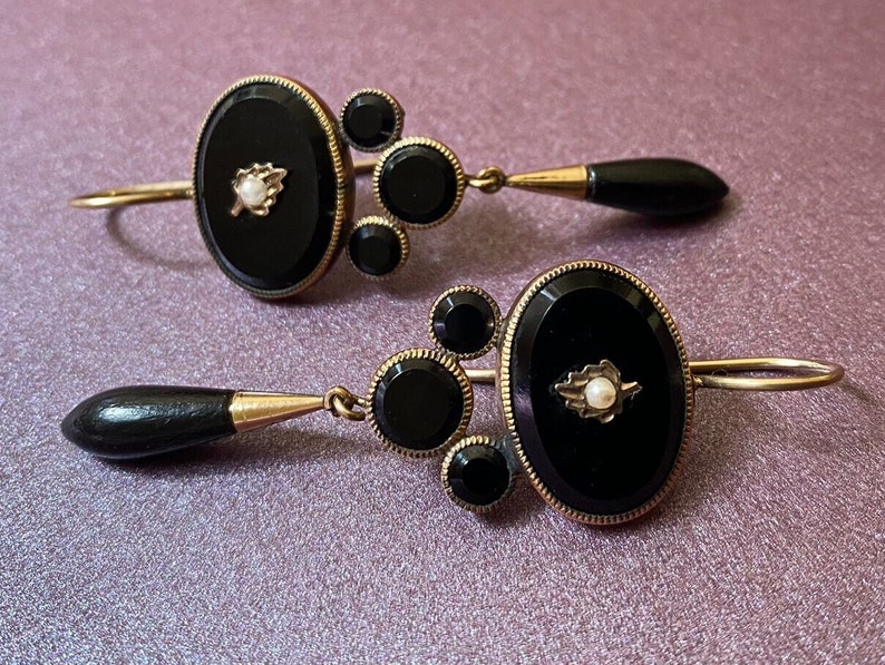May include: A pair of black and gold earrings with a teardrop-shaped black stone dangle. Each earring features a round black stone with a smaller black stone on either side. The center stone is surrounded by a gold setting with a small pearl in the center.
