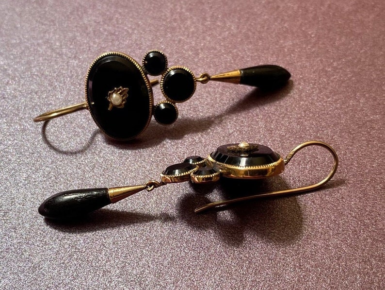 May include: A pair of antique earrings with black onyx and pearl accents. The earrings are made of gold and have a dangling design.