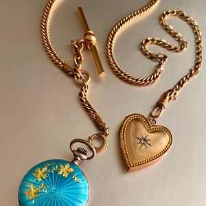 May include: A gold chain with a T-bar, a round pocket watch with a bright blue guilloch&eacute; enamel finish featuring gold leaf floral accents, and a heart-shaped gold locket with a central star-set diamond, all connected by a metal link chain.