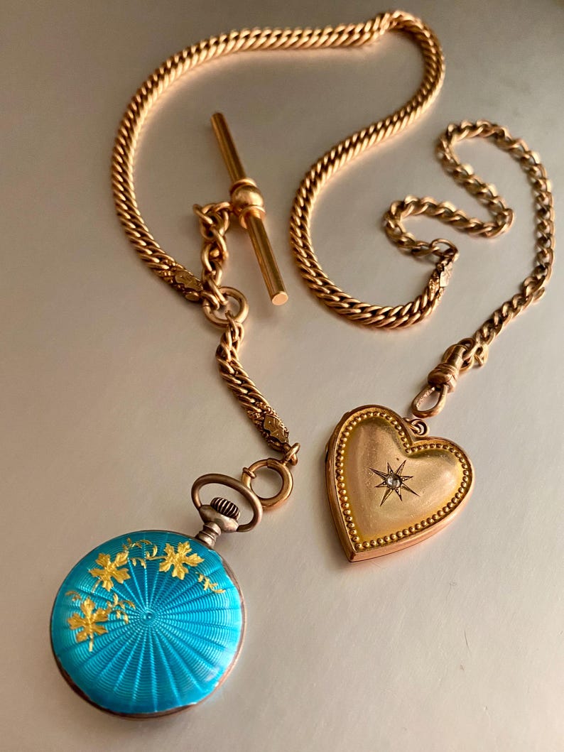 May include: A gold chain with a T-bar, a round pocket watch with a bright blue guilloch&eacute; enamel finish featuring gold leaf floral accents, and a heart-shaped gold locket with a central star-set diamond, all connected by a metal link chain.