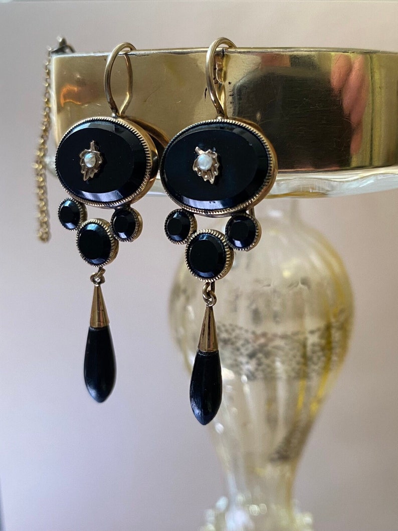May include: A pair of antique black onyx drop earrings with gold accents and a pearl detail. The earrings feature a teardrop-shaped pendant and a cluster of round stones.