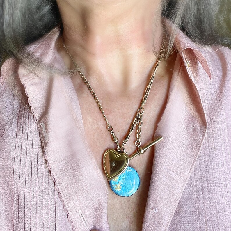 May include: A gold-toned necklace featuring a heart-shaped locket and a round, bright blue enamel pendant with a starburst pattern. The necklace includes a T-bar toggle clasp and a delicate chain, resting against a light pink button-down shirt.