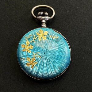May include: A round pocket watch with a bright turquoise guilloch&eacute; enamel finish featuring a radiating sunburst pattern. The surface is decorated with gold-toned metallic leaf motifs on the left side and includes a silver-toned winding crown.