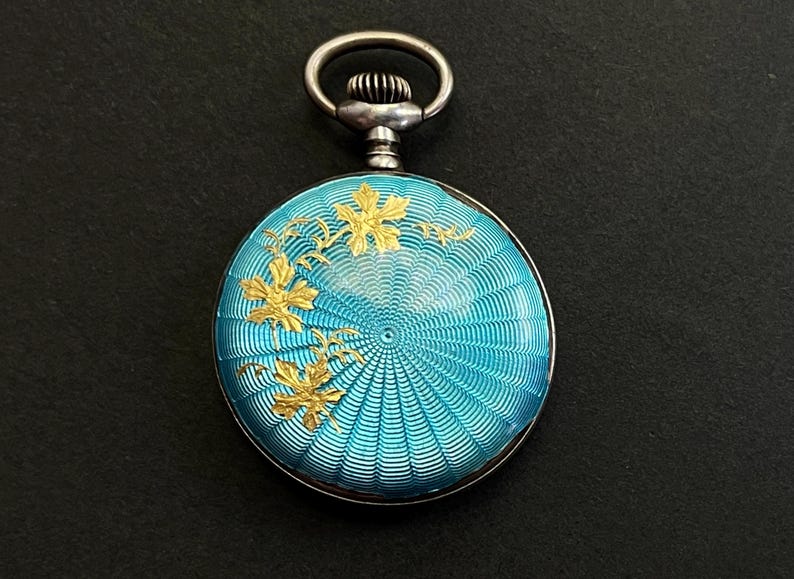 May include: A round pocket watch with a bright turquoise guilloch&eacute; enamel finish featuring a radiating sunburst pattern. The surface is decorated with gold-toned metallic leaf motifs on the left side and includes a silver-toned winding crown.