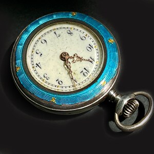 May include: A round pocket watch featuring a bright turquoise blue guilloch&eacute; enamel bezel with small gold star accents. The watch face is off-white with black Arabic numerals and ornate gold-toned hands. A silver-toned winding crown and bow are attached.