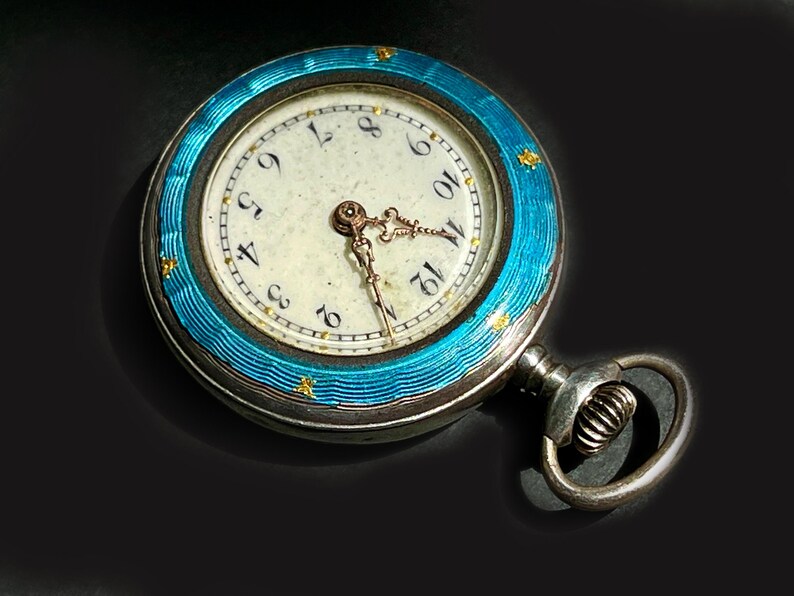 May include: A round pocket watch featuring a bright turquoise blue guilloch&eacute; enamel bezel with small gold star accents. The watch face is off-white with black Arabic numerals and ornate gold-toned hands. A silver-toned winding crown and bow are attached.
