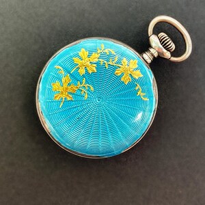 May include: A round pocket watch featuring a vibrant turquoise guilloch&eacute; enamel finish with a radiating sunburst pattern. The surface is decorated with delicate gold-toned leaf motifs. It includes a silver-toned winding crown and a circular bow.