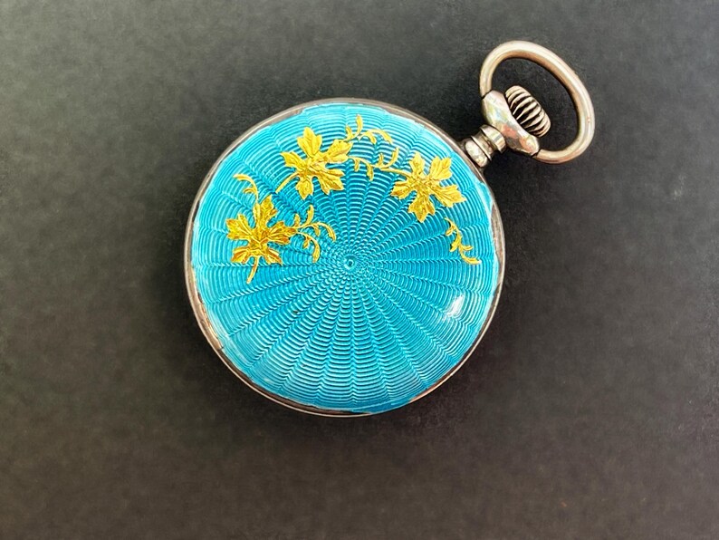 May include: A round pocket watch featuring a vibrant turquoise guilloch&eacute; enamel finish with a radiating sunburst pattern. The surface is decorated with delicate gold-toned leaf motifs. It includes a silver-toned winding crown and a circular bow.