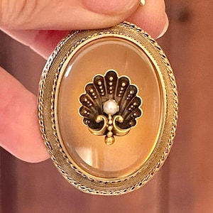 May include: An oval gold brooch featuring a textured metal border and a smooth, translucent amber-colored center. The center displays a dark, fan-shaped shell motif with a small, round white pearl at its base, set against the warm, light brown background.