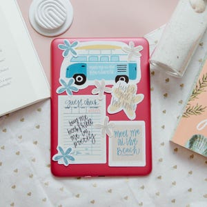 May include: A pink e-reader with stickers, an open book, a book titled "It's Not Summer Without You", a small glass jar with a cork, and a white decorative item are arranged on a patterned cloth.