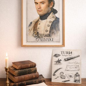 Puede incluir: Un retrato enmarcado de Benjamin Tallmadge, una figura histórica, cuelga sobre una colección de libros y documentos antiguos. Una vela encendida en un candelabro de latón y un póster de Turn con ilustraciones completan la escena.