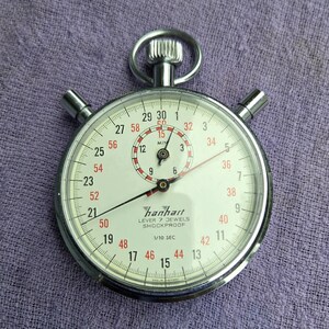 May include: A silver-toned Hanhart stopwatch with a white face, black and red numerals, and two pushers. The dial features the text Hanhart, LEVER 7 JEWELS, SHOCKPROOF, and 1/10 SEC. A small sub-dial indicates minutes, and a crown sits at the top.