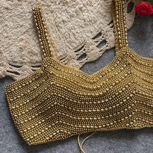 May include: A gold beaded top with a V-neck and thin straps. The surface is covered in small, round beads, creating a textured effect. The straps and the bottom of the top have a simple design. The top is a neutral color, suitable for various outfits.