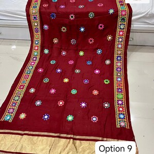May include: A maroon textile with intricate embroidery and mirror work. The fabric features colorful circular embellishments and a decorative border with geometric patterns. The bottom edge has a gold-colored trim. The text "Option 9" is visible.