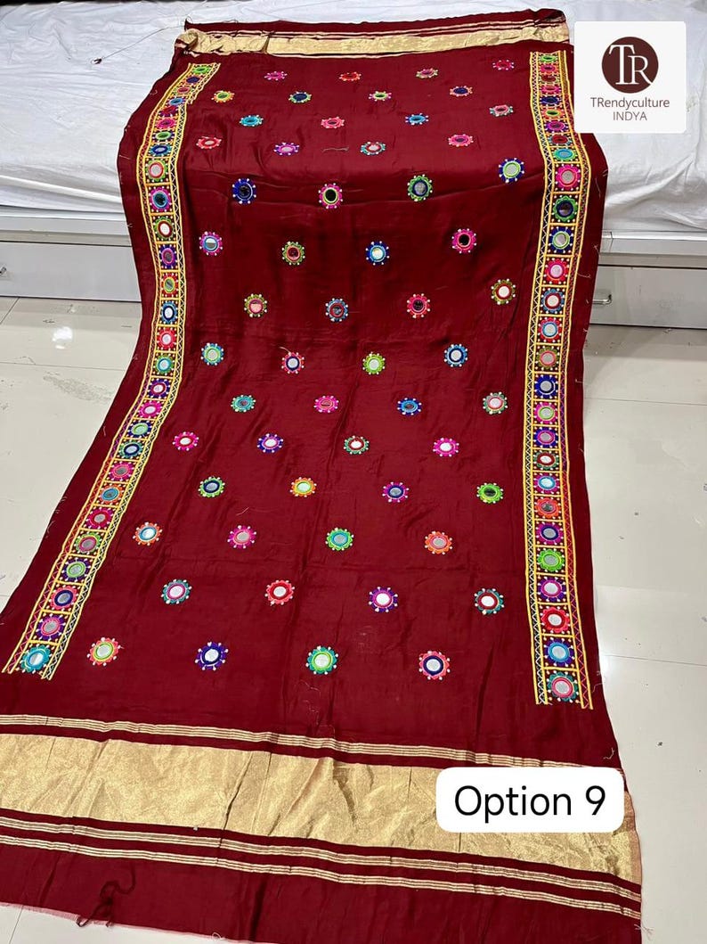 May include: A maroon textile with intricate embroidery and mirror work. The fabric features colorful circular embellishments and a decorative border with geometric patterns. The bottom edge has a gold-colored trim. The text "Option 9" is visible.