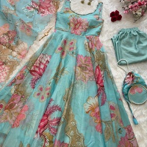 Op de afbeelding: Een lichtblauwe jurk met bloemenprint met bijpassende broek, sjaal en een kleine handtas. De jurk heeft een ronde hals en is versierd met roze en bruine bloempatronen. De set bevat een paar gouden oorbellen en een parelketting.