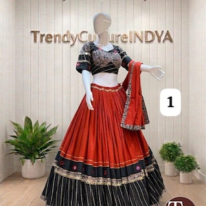 May include: A two-piece lehenga set featuring a black short-sleeved blouse with white floral embroidery and a long, pleated red skirt. The skirt has a wide black hem with gold vertical stripes and circular mirror accents. A matching red dupatta is included.