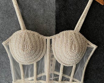 Può includere: Top bustier color avorio con abbellimenti di perle. Il top presenta un corpetto strutturato con un rivestimento in rete trasparente e delicati dettagli di perle sulle spalline e sulle cuciture. Il testo "Opzione 1" è visibile.