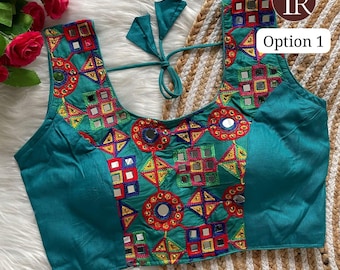 May include: Teal embroidered top with colourful geometric patterns and small mirror accents. The top has a scoop neckline and tie closure. The text "Option 1" is visible on the upper right corner. The top is displayed on a white and tan surface.