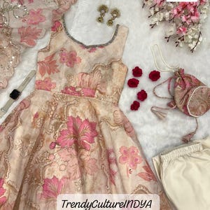 Könnte beinhalten: Beige-rosa Kleid mit floralem Muster, passendem Schal, Ohrringen und kleiner Handtasche. Das Kleid hat ein ärmelloses Design und einen dekorativen Ausschnitt. Der Text "TrendyCultureINDYA" ist auf dem Kleid zu sehen.