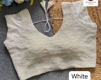 Può includere: Un top bianco senza maniche con scollo a V profondo, riccamente decorato con piccole perline rotonde simili a perle. L'intricato lavoro di perline forma un motivo a conchiglia sul davanti. La parola "White" è visualizzata in un rettangolo bianco in basso.