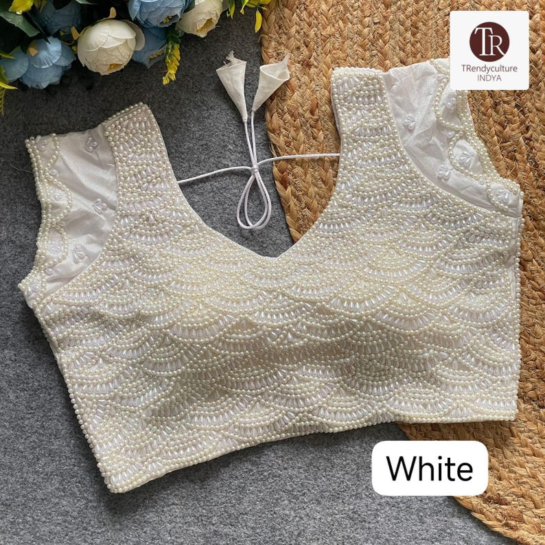 May include: A white, sleeveless top with a deep V-neck, adorned with numerous small, round, pearl-like beads. The beadwork creates a scalloped design across the front. The word "White" is displayed in a white rectangle at the bottom.