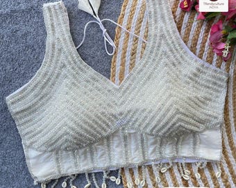 Può includere: Top senza maniche bianco con perline con scollo a V. La parte superiore è pesantemente impreziosita da perline d'argento con un motivo a spina di pesce. L'orlo inferiore presenta accenti di conchiglie pendenti. Il testo "Opzione 1" è visibile.