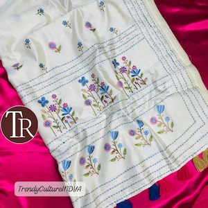 May include: White Assam cotton fabric with blue, pink, and purple floral embroidery. The fabric has a border with blue stitching and tassels in gold, blue, and red. The text "Assam cotton" and "TrendyCultureINDYA" are visible.