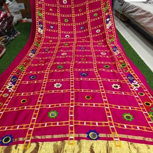 May include: A vibrant pink textile with a grid pattern, adorned with embroidered flowers and small mirrors. The fabric has a gold border and trim. The text "Option 1" is visible in the lower right corner. The textile is likely a decorative item.