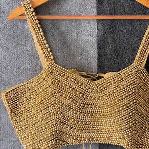 May include: A gold-coloured beaded top with a scoop neckline and spaghetti straps. The top is densely covered in small, round beads, creating a textured surface. It hangs on a wooden hanger against a grey and black background.