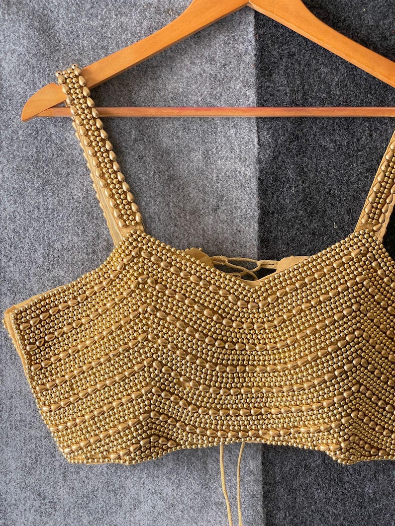 May include: A gold-coloured beaded top with a scoop neckline and spaghetti straps. The top is densely covered in small, round beads, creating a textured surface. It hangs on a wooden hanger against a grey and black background.