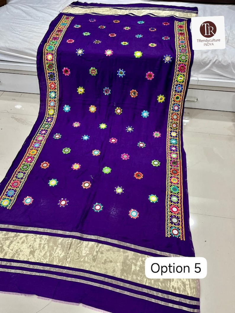 May include: A vibrant purple textile with intricate embroidery and mirror work. The edges feature a detailed border with colorful geometric patterns. Scattered across the fabric are small, circular mirrors, adding a reflective quality. The bottom edge has a gold metallic trim. The text "Option 5" is visible.
