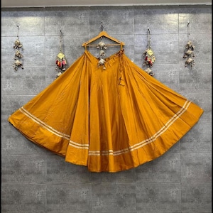 Könnte beinhalten: Senfgelber Rock mit weitem, fließendem Design. Der Rock hat zwei horizontale weiße Streifen am Saum. Der Rock hängt an einem Holzbügel vor einer grau gefliesten Wand.