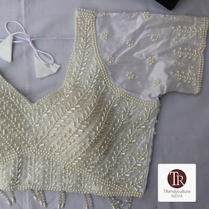 Puede incluir: Blusa color marfil con cuentas y perlas, con escote en V y mangas cortas. El corpiño está profusamente adornado con cuentas de semillas y perlas en un patrón de hojas. Las mangas tienen detalles de perlas. La blusa tiene cierre de lazo y un logo en la esquina inferior derecha.