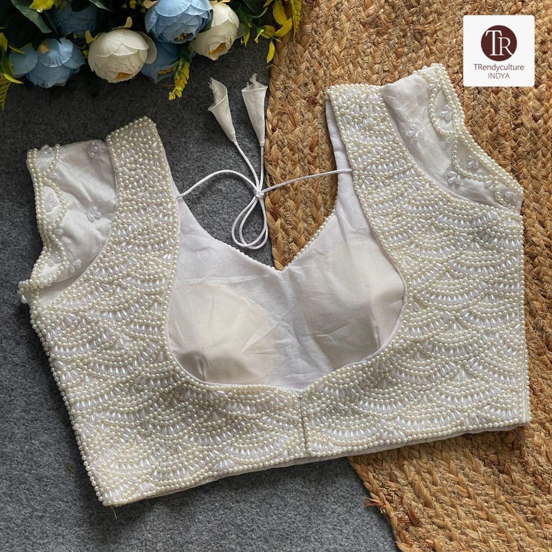 May include: A white, sleeveless blouse adorned with pearl beadwork. The blouse has a deep V-neck and a tie closure at the back. The pearl embellishments create a textured design across the bodice and neckline, adding a touch of elegance to the garment.