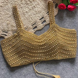 Pode incluir: Top dourado com contas, decote em V e alças finas. A parte superior é densamente coberta por pequenas contas redondas, criando uma superfície texturizada. Duas borlas pendem da parte inferior. É provavelmente uma peça de roupa formal ou festiva.
