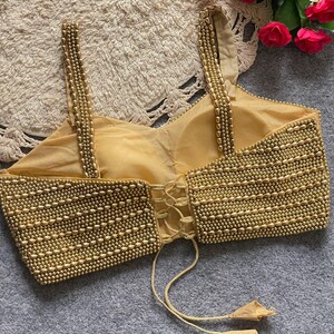 May include: A gold-coloured, beaded top with spaghetti straps and a corset-style back. The bodice is heavily embellished with small, round beads, creating a textured surface. The straps are also adorned with beads. The top has a tie closure with tassels.