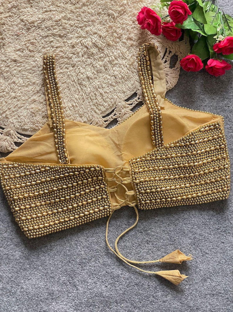 May include: A gold-coloured, beaded top with spaghetti straps and a corset-style back. The bodice is heavily embellished with small, round beads, creating a textured surface. The straps are also adorned with beads. The top has a tie closure with tassels.