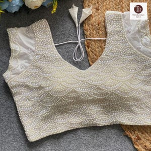 May include: Cream-coloured, sleeveless blouse with a deep V-neck. The bodice is heavily embellished with small, round, pearl-like beads arranged in a scalloped pattern. The shoulders and upper back feature delicate, sheer fabric. The blouse has a tie closure.