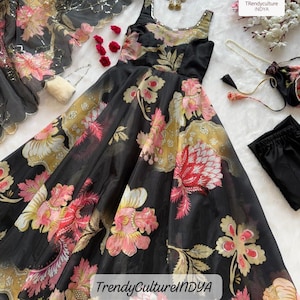 May include: Black sleeveless dress with a floral pattern in pink, yellow, and red. The dress has a scoop neckline and a flowing skirt. A matching scarf and accessories are also visible. The text "TrendyCultureINDYA" is printed on the dress.