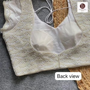 May include: A white, sleeveless top featuring detailed pearl beading. The neckline and edges are decorated with small, round pearls. The back view of the top is shown, with a tie closure. The text "Back view" is visible at the bottom.