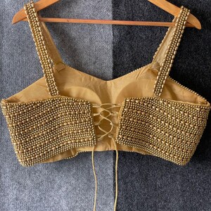 May include: A gold-coloured, beaded top with spaghetti straps and a lace-up back. The bodice and straps are heavily embellished with small, round beads. The top is hanging on a wooden hanger against a grey and black background.