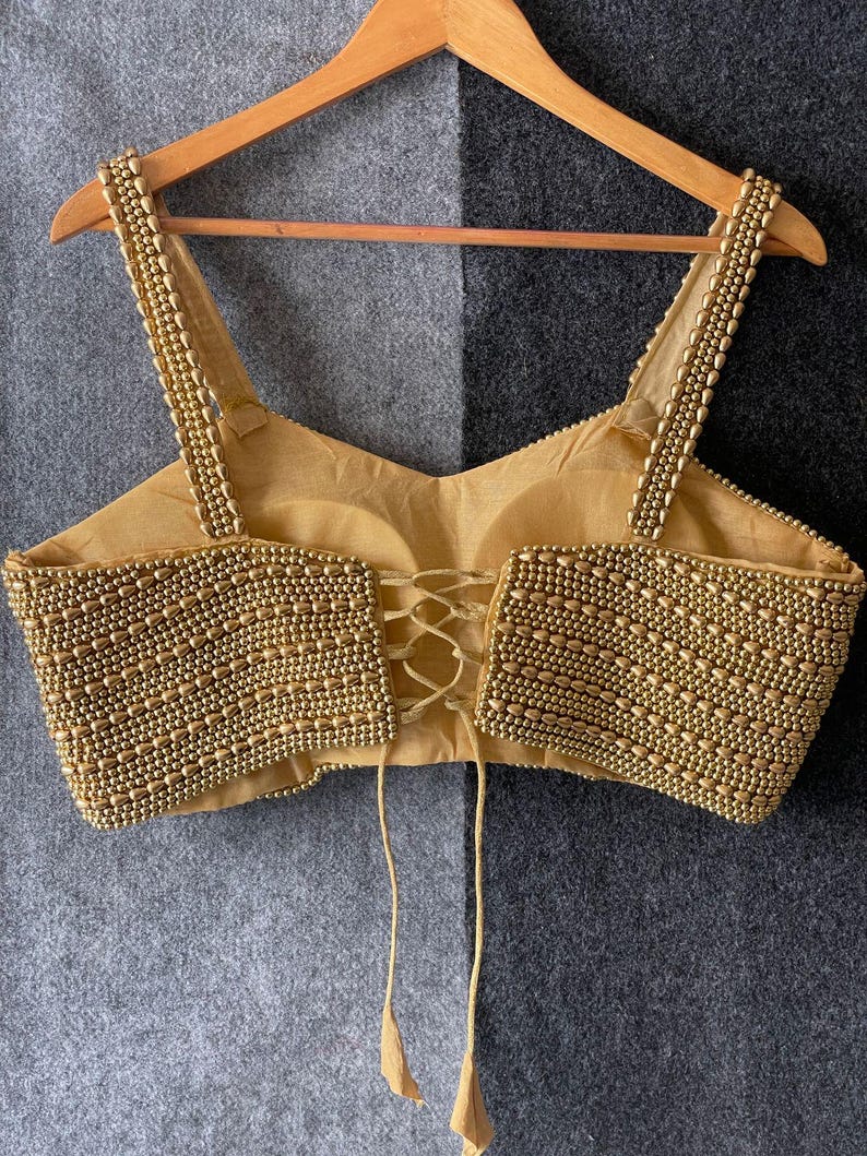 May include: A gold-coloured, beaded top with spaghetti straps and a lace-up back. The bodice and straps are heavily embellished with small, round beads. The top is hanging on a wooden hanger against a grey and black background.