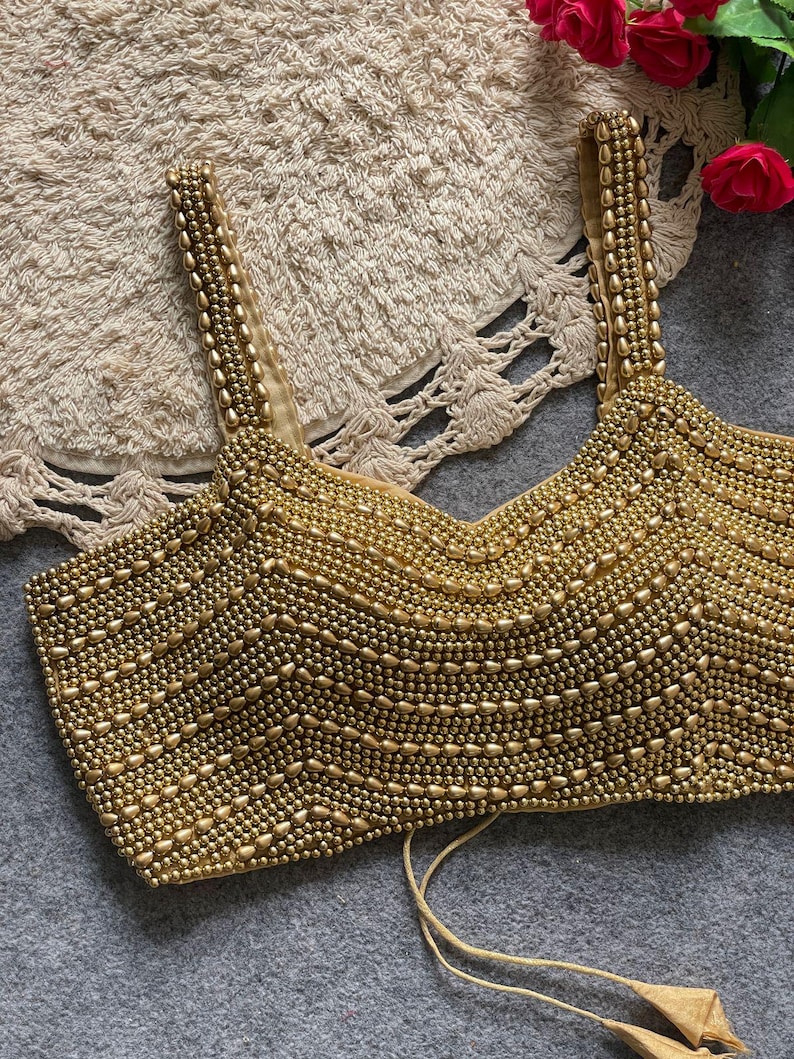 May include: A gold beaded top with a V-neck and thin straps. The surface is covered in small, round beads, creating a textured effect. The straps and the bottom of the top have a simple design. The top is a neutral color, suitable for various outfits.